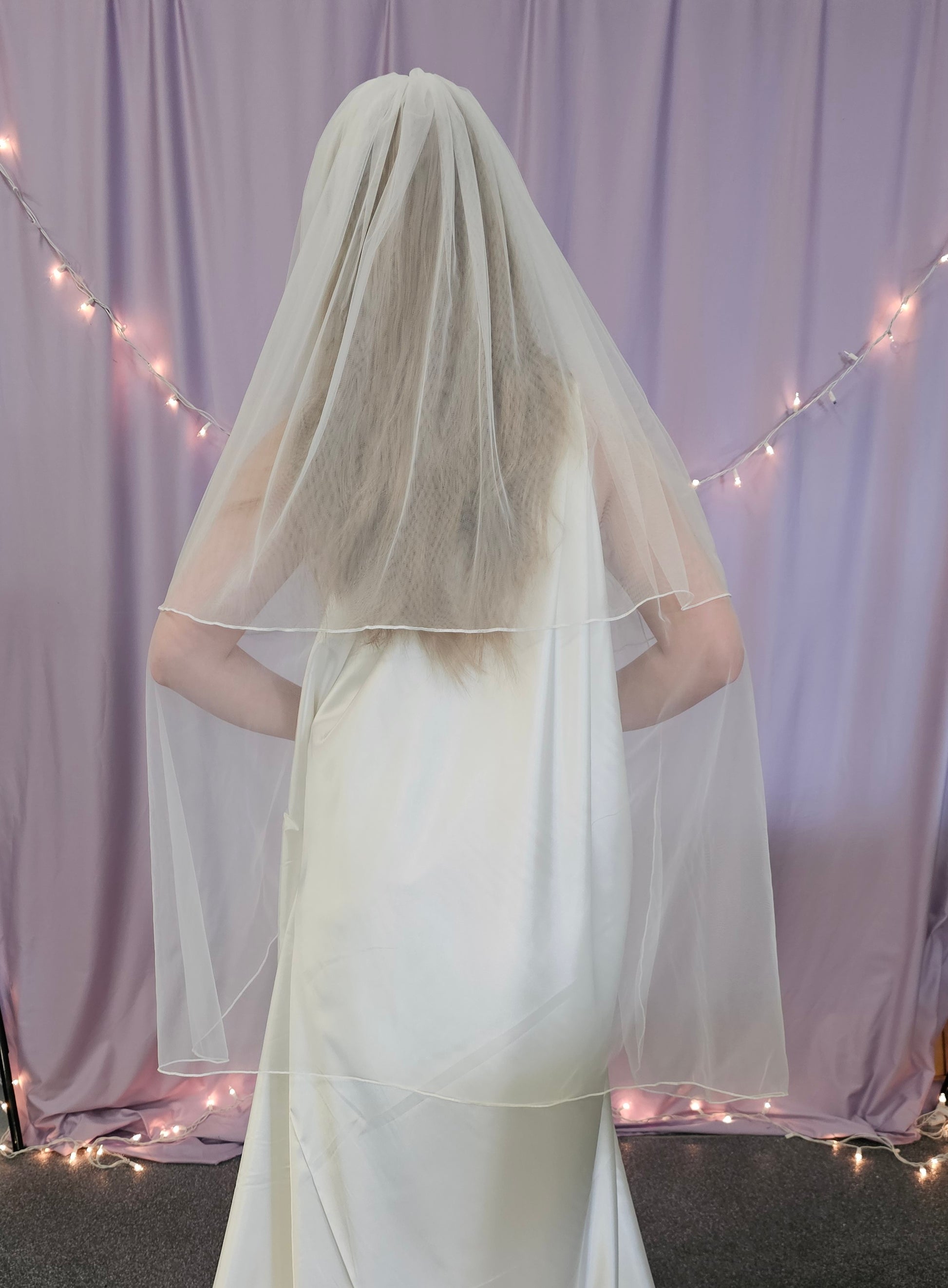 Person wearing a white wedding dress with a long veil in front of a light purple curtain with string lights.