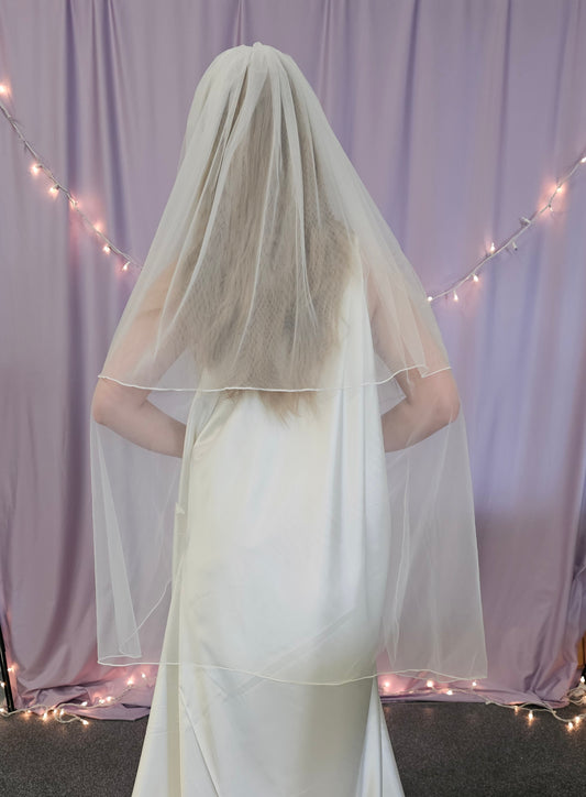 Person wearing a white wedding dress with a long veil in front of a light purple curtain with string lights.
